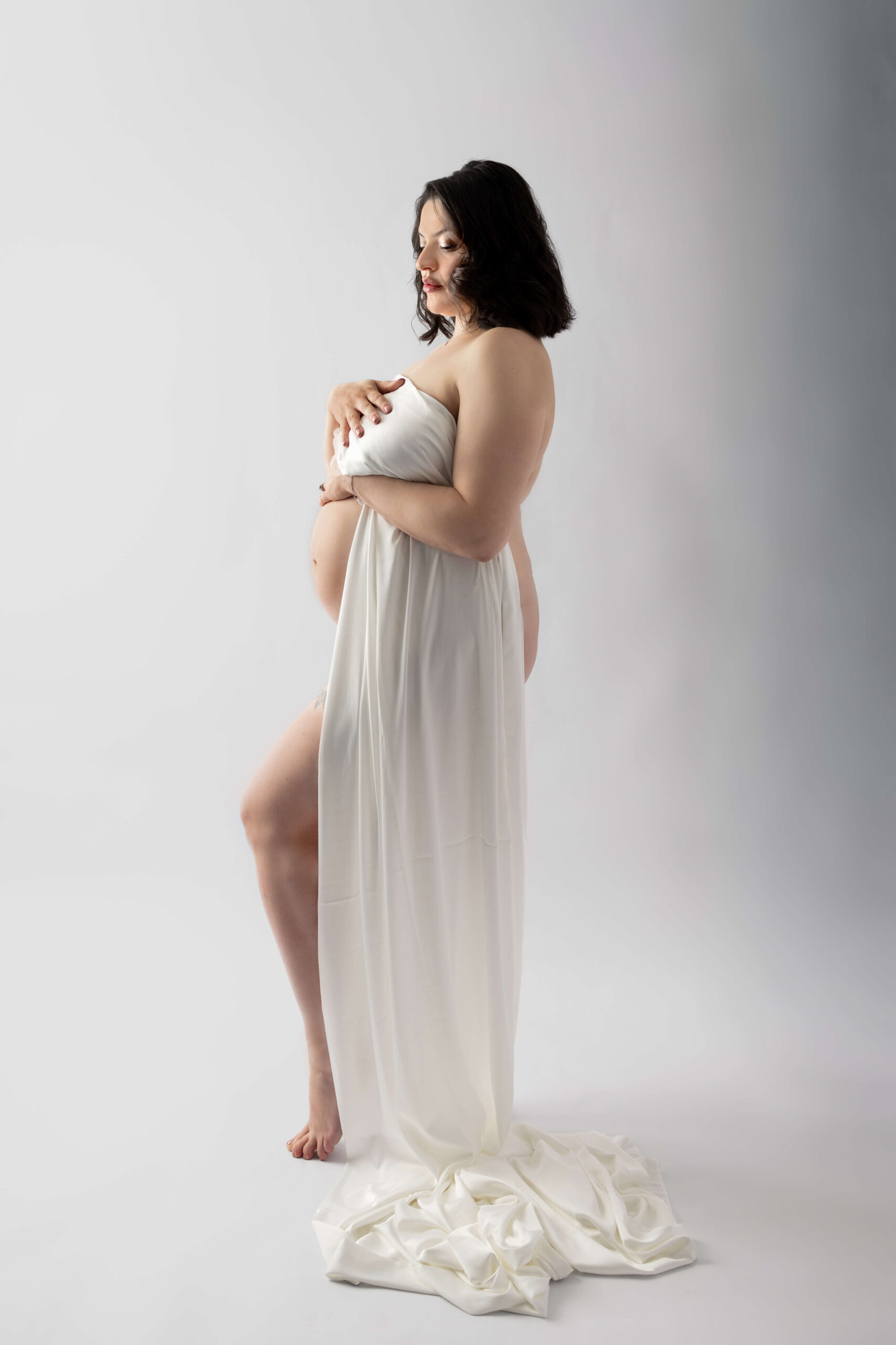 A pregnant woman stands with a knee popped while wrapped in a white silk in a studio
