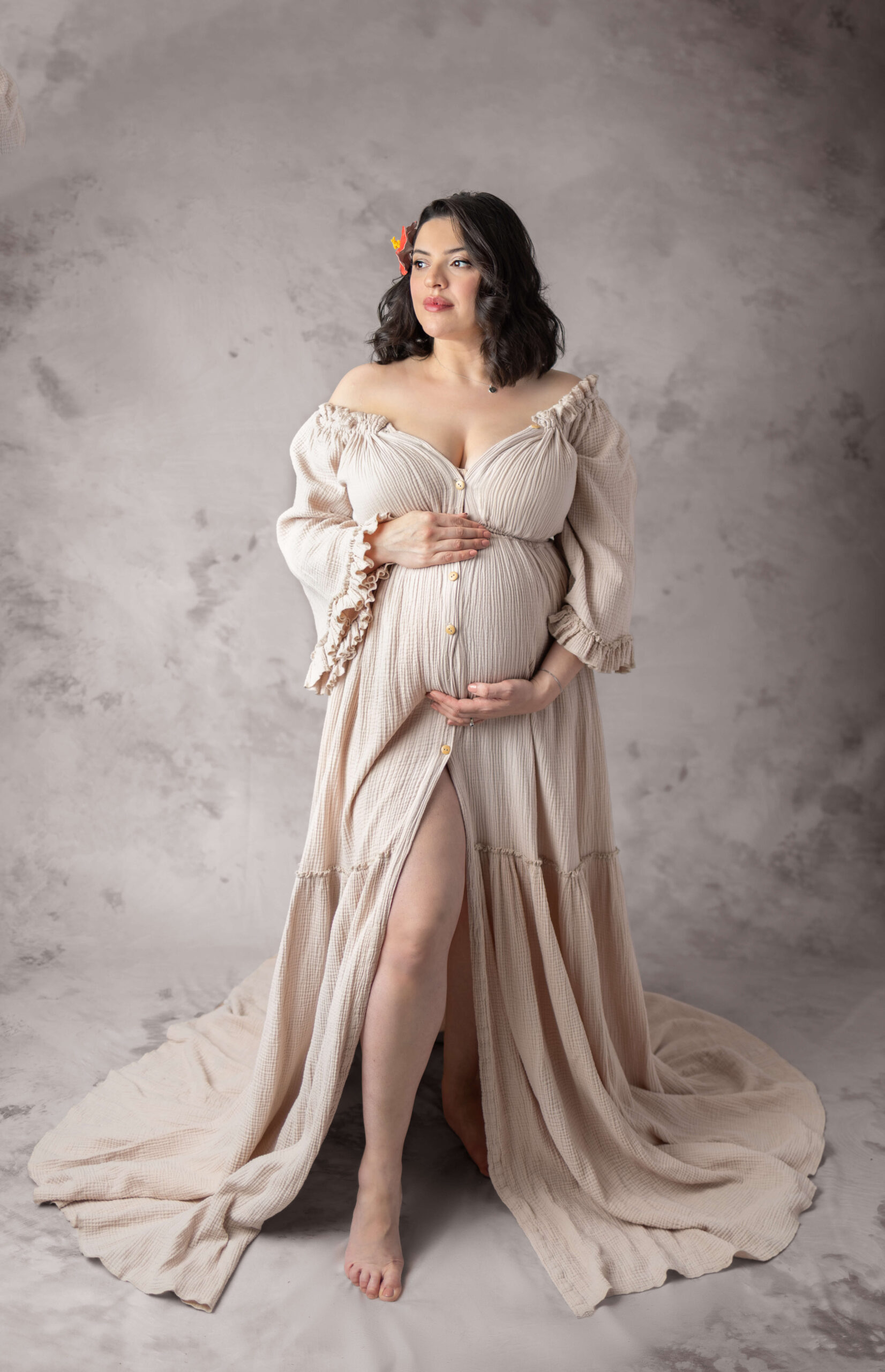 A pregnant woman stands in a cream maternity gown in a studio holding her bump and looking into the distance after her 3d ultrasound in bergen county nj