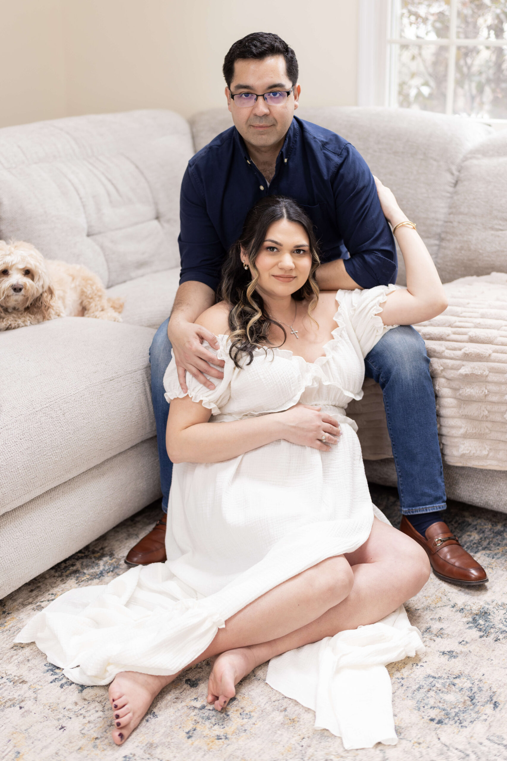 An expectant woman sits on the floor leaning into the lap of her husband on the couch in a blue shirt