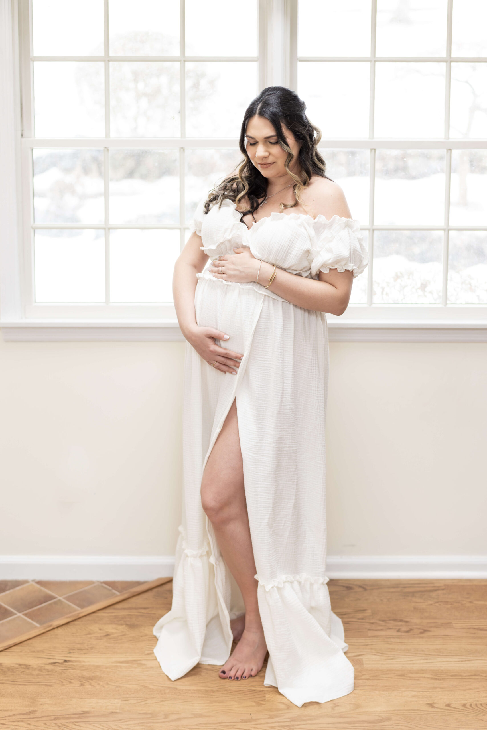 A mother to be in a white maternity gown smiles down to her bump with hands on it after meeting with doulas in bergen county nj