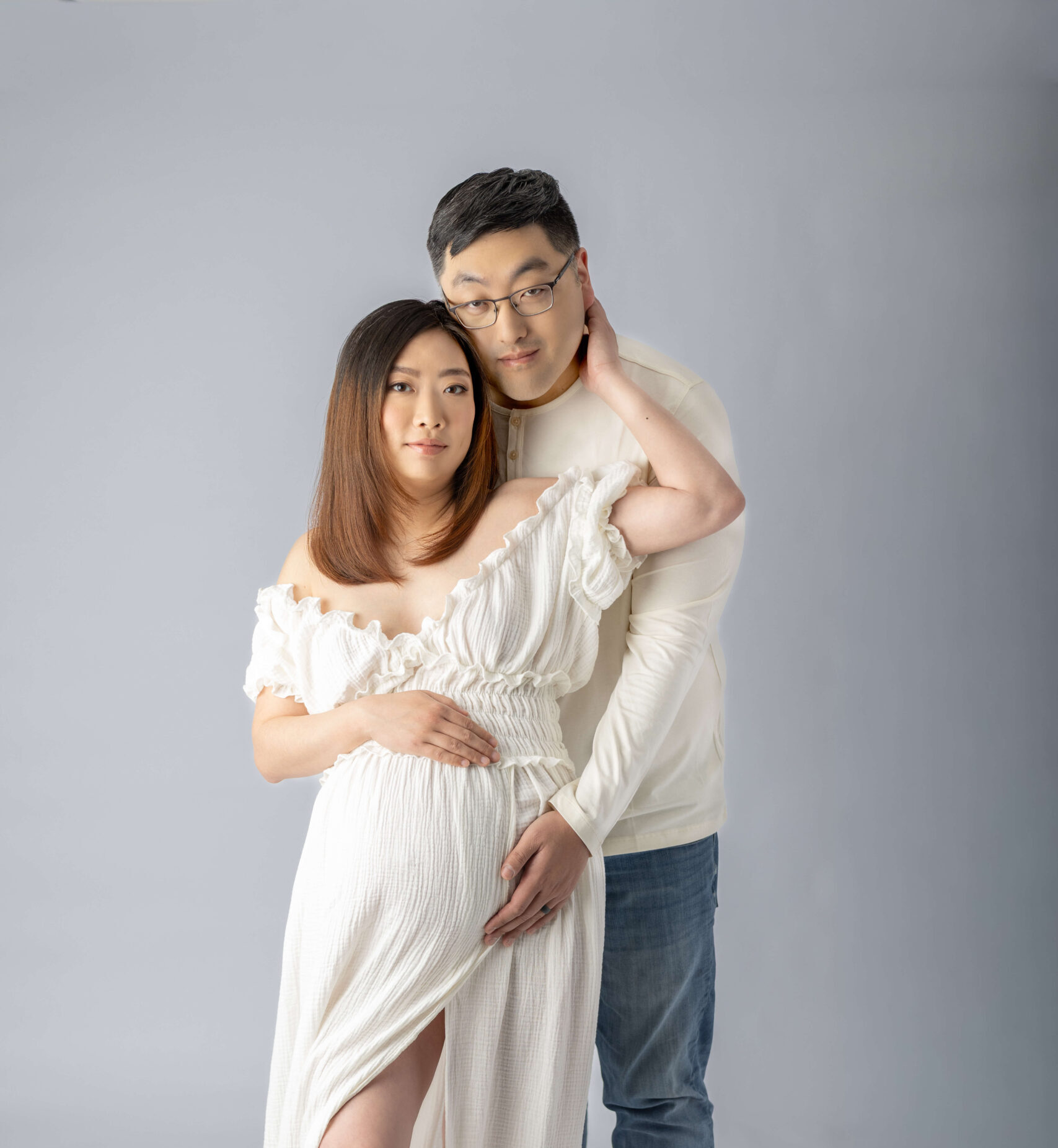 A happy couple in white snuggle while standing in a studio with hands on the bump