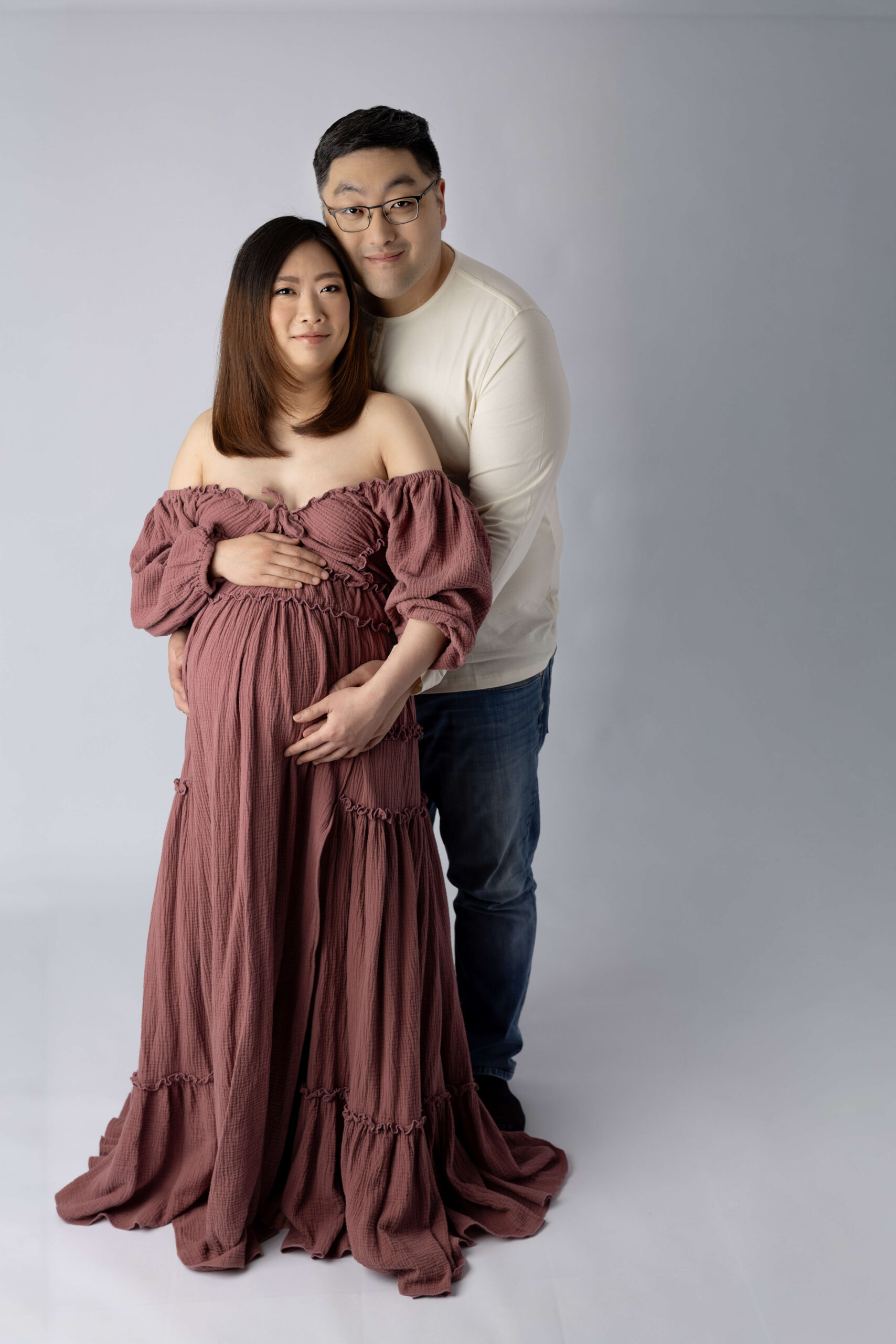 A mom and dad stand in a studio smiling with hands on the bump in a cream shirt and long purple maternity gown after visiting our birthing center morristown