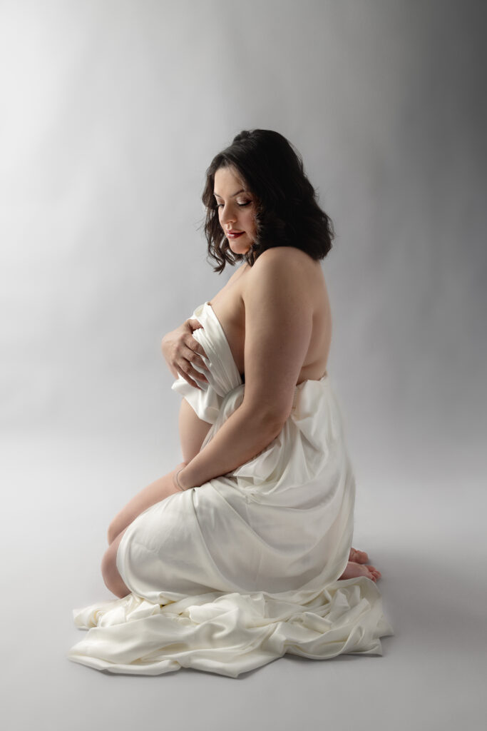 Maternity portrait of expectant mother draped in flowing white fabric, posed against a soft studio backdrop.