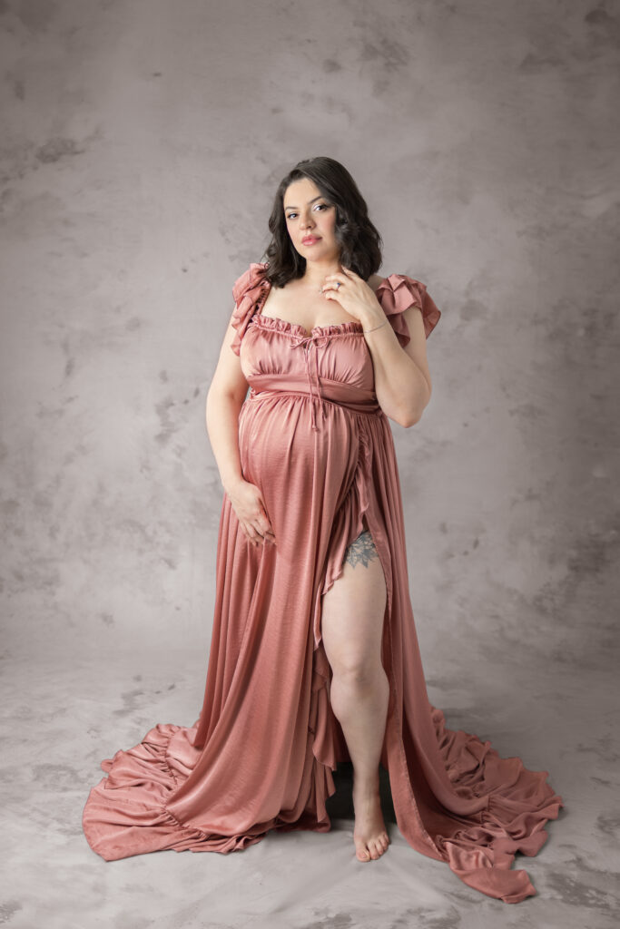 Mother-to-be poses in a rose gown with one hand on her belly against a textured backdrop.