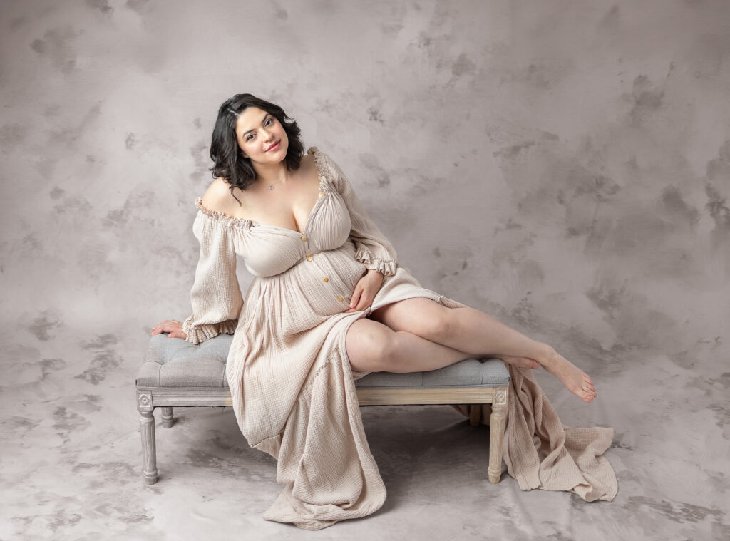 Smiling expectant mother reclines on a vintage bench in a cream gown against a soft textured backdrop.