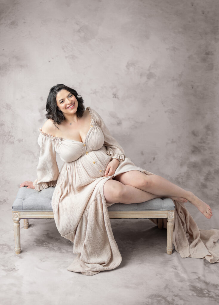Smiling expectant mother reclines on a vintage bench in a soft beige gown in Clifton, NJ.