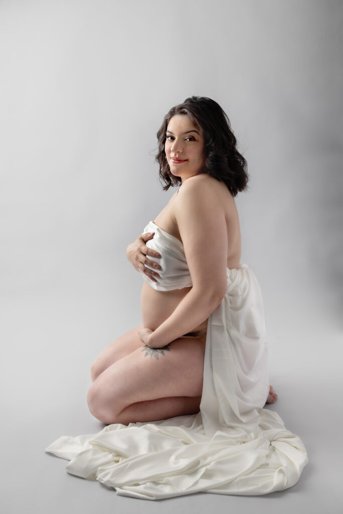 Expectant mother kneels in a white draped fabric, gently holding her belly and looking toward the camera.
