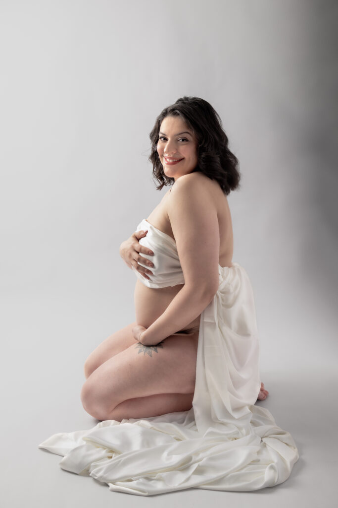Smiling mother-to-be kneels in white fabric, cradling her belly during a Clifton, NJ maternity session.