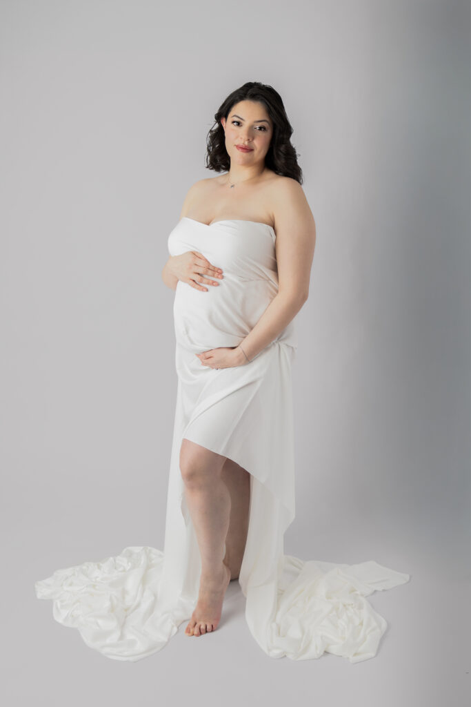 Expectant mother stands in a white draped gown, gently cradling her belly against a simple backdrop.