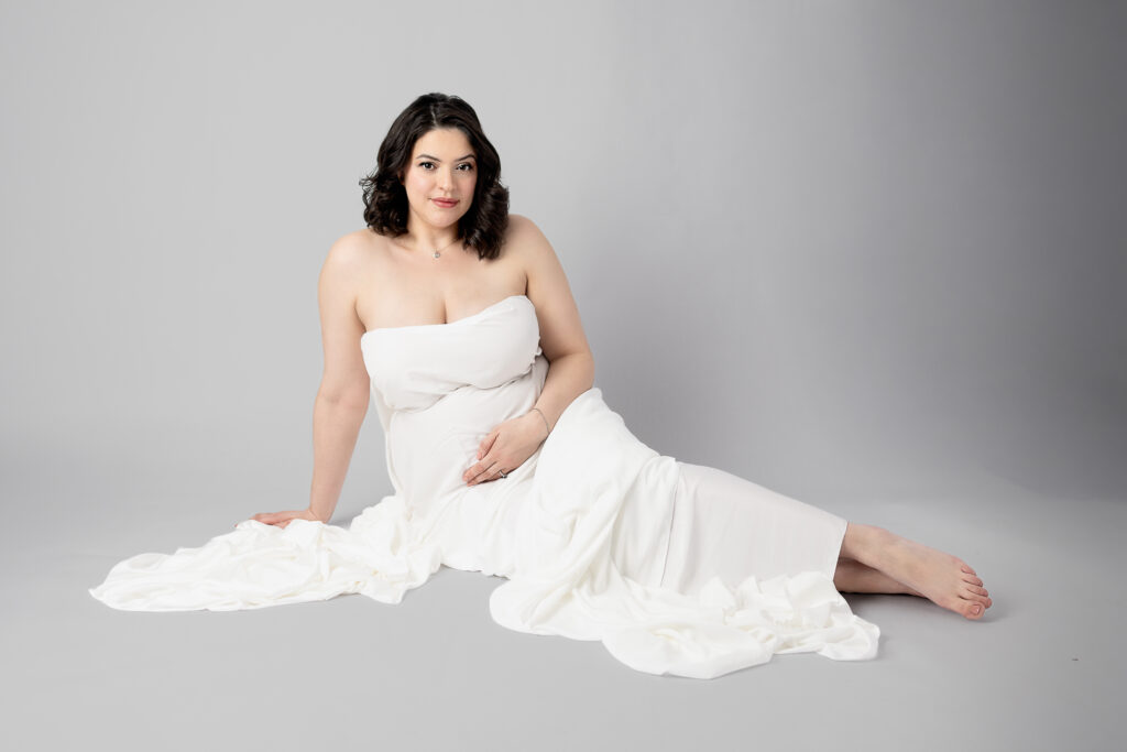 Mother-to-be reclines in a white gown, surrounded by flowing fabric during a Clifton, NJ maternity portrait.