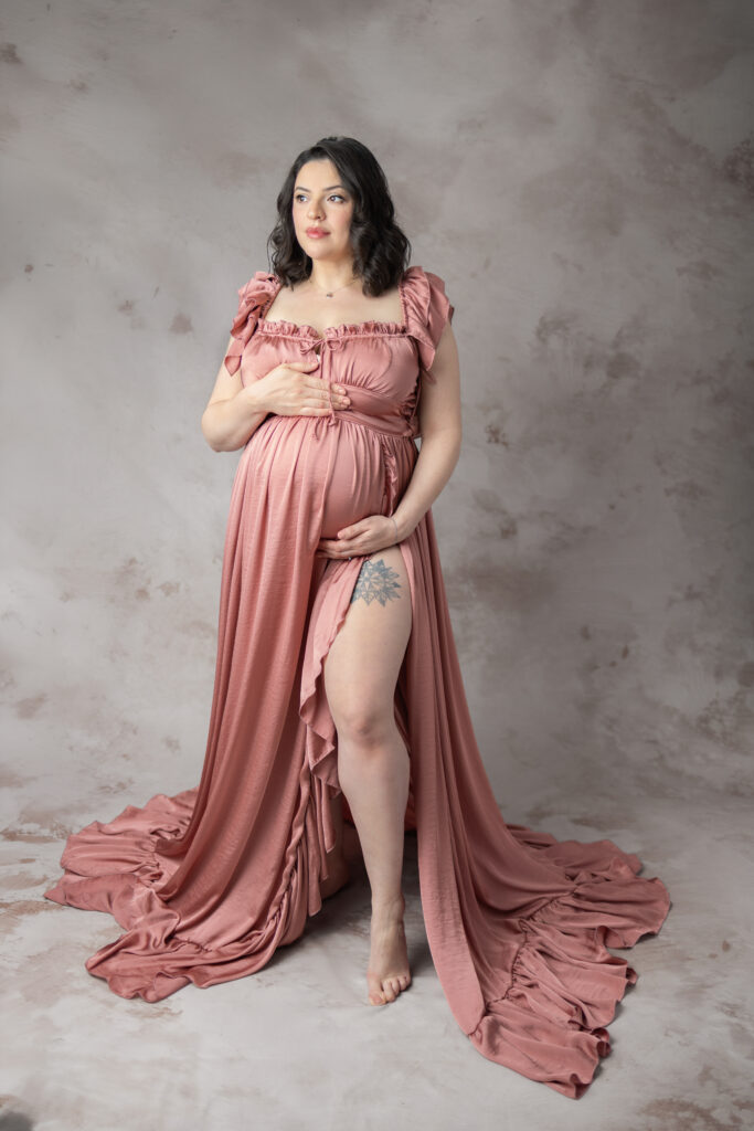Pregnant mother poses in a rose-colored gown, looking away against a softly textured studio backdrop.