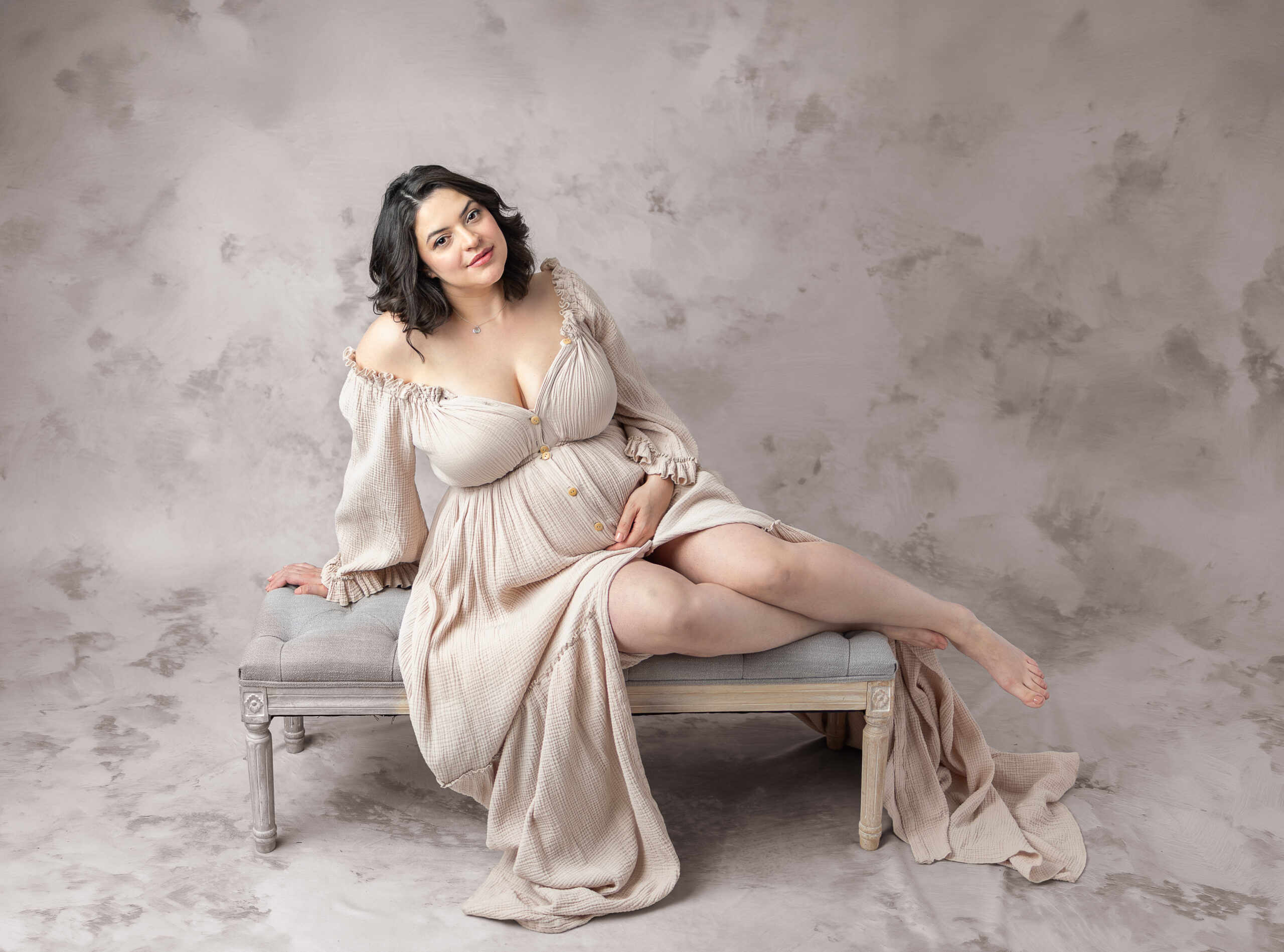 A mother to be in a beige maternity gown sitting in a studio with a hand on her belly after enjoying her 3d ultrasound in bergen county nj