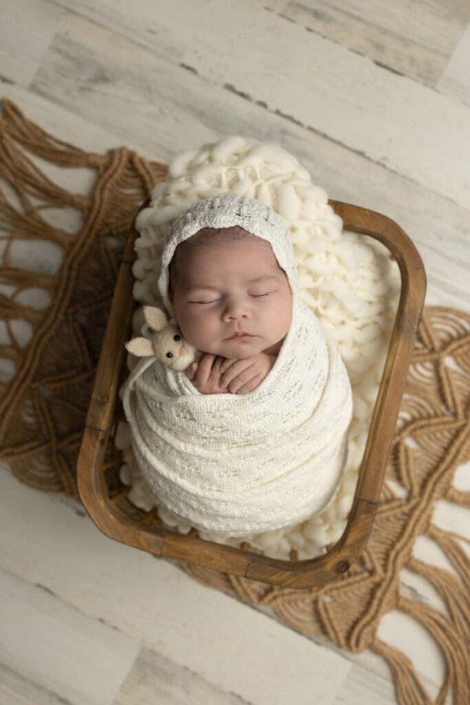 Newborn baby girl sleeping peacefully in a wooden crate with a cream wrap, knit bonnet, and small bunny prop during a posed newborn photography session.