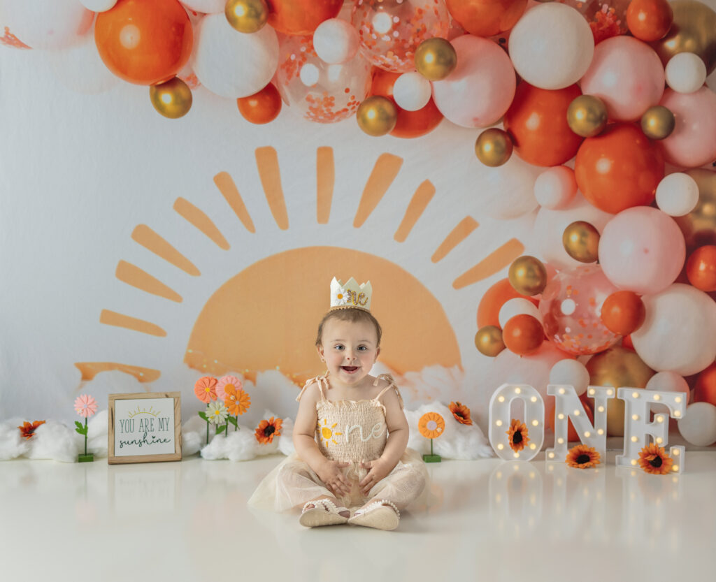 Smiling one-year-old sits beneath a orange sunshine backdrop and balloon garland during a joyful cake smash session.