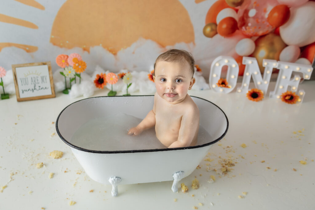Smiling birthday girl relaxes in a white tub after her cake smash session.