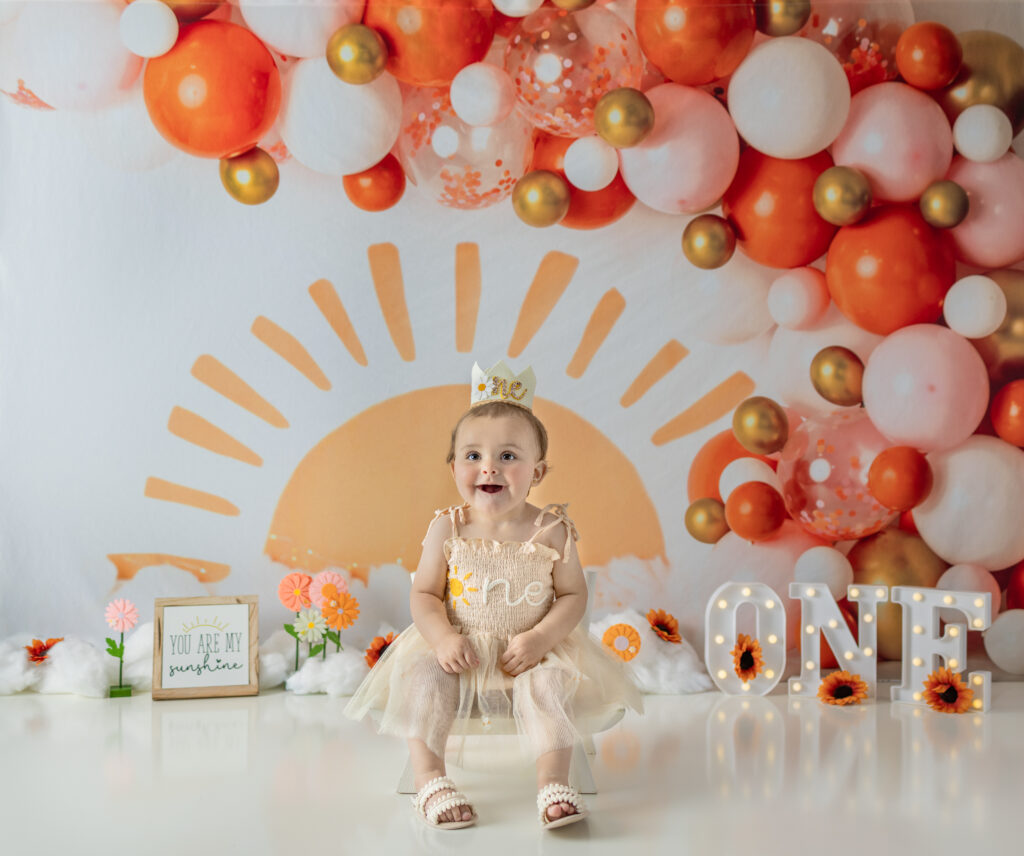 One-year-old girl sits on a small chair beneath balloon garland during a Bergen County cake smash session.