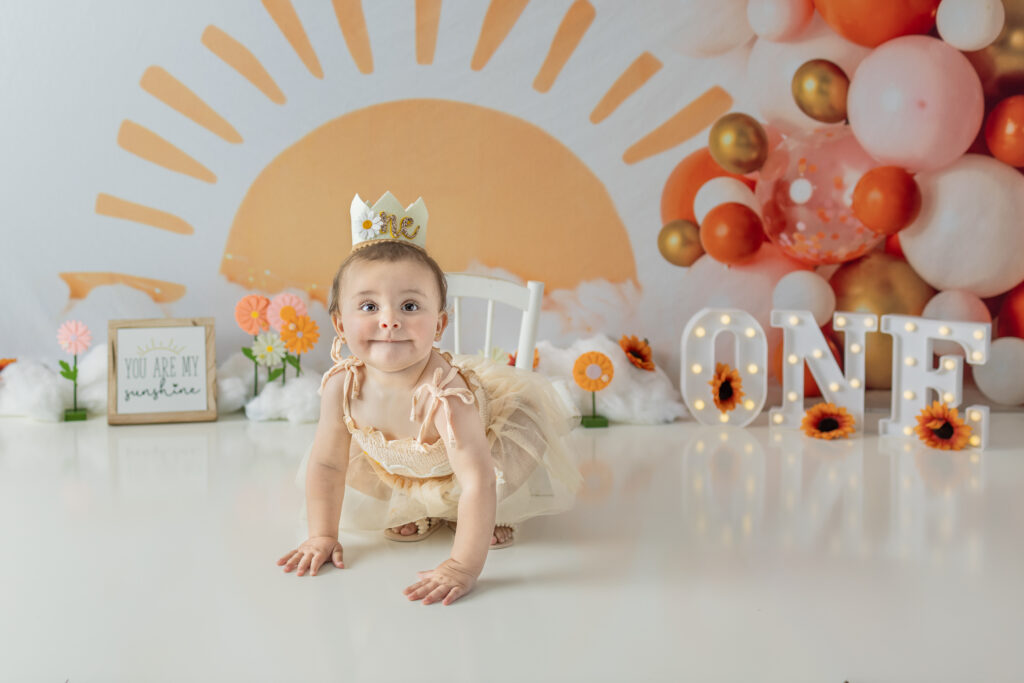 One-year-old girl crawls toward camera during playful sunshine-themed cake smash session.