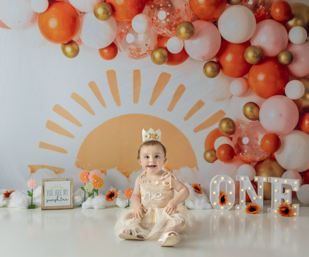 One-year-old girl smiles beneath orange and pink balloons during a Bergen County cake smash session.