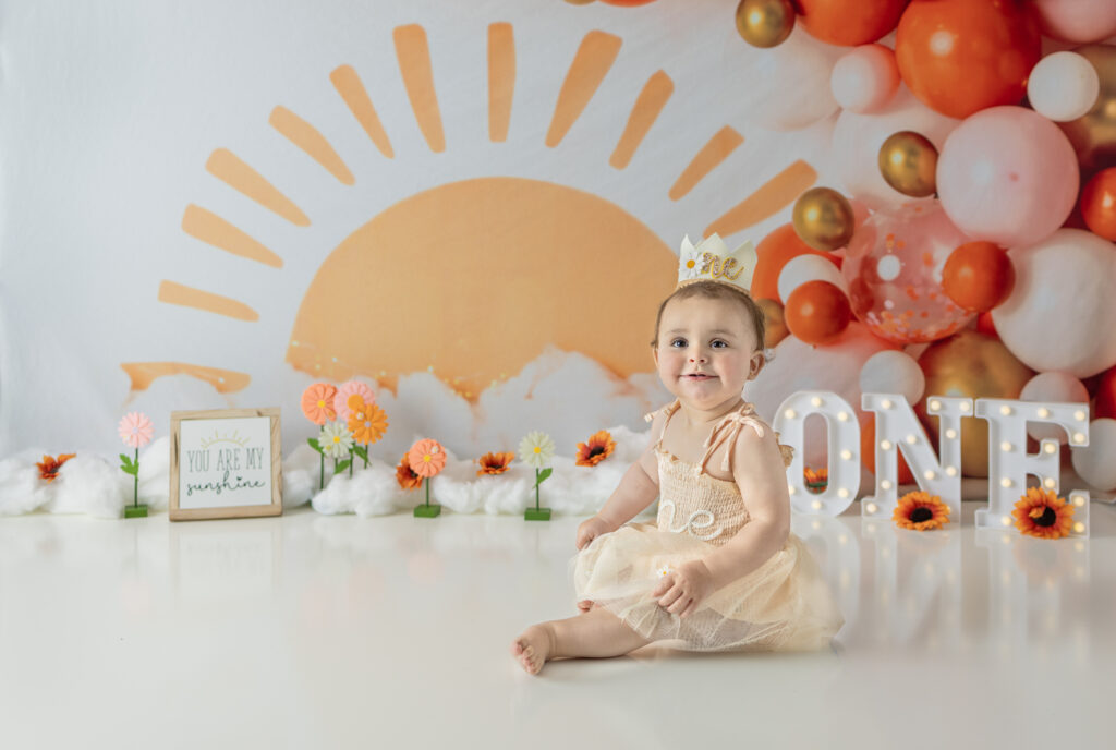 Birthday girl sits beside illuminated ONE letters during a sunshine-themed first birthday session.