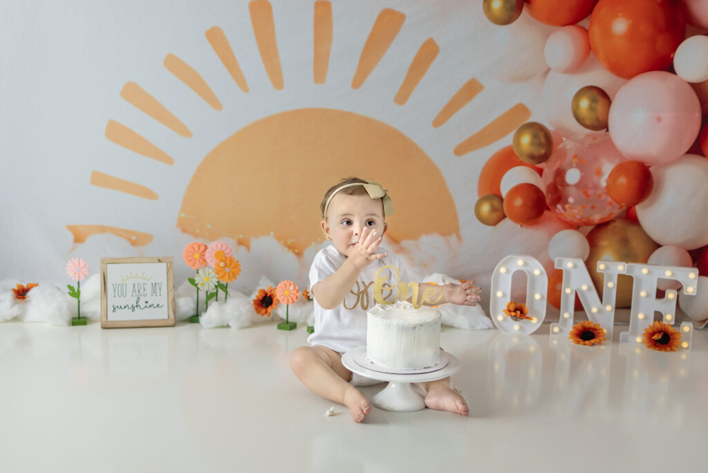 One-year-old girl tastes frosting from birthday cake during sunshine-themed cake smash in Bergen County.