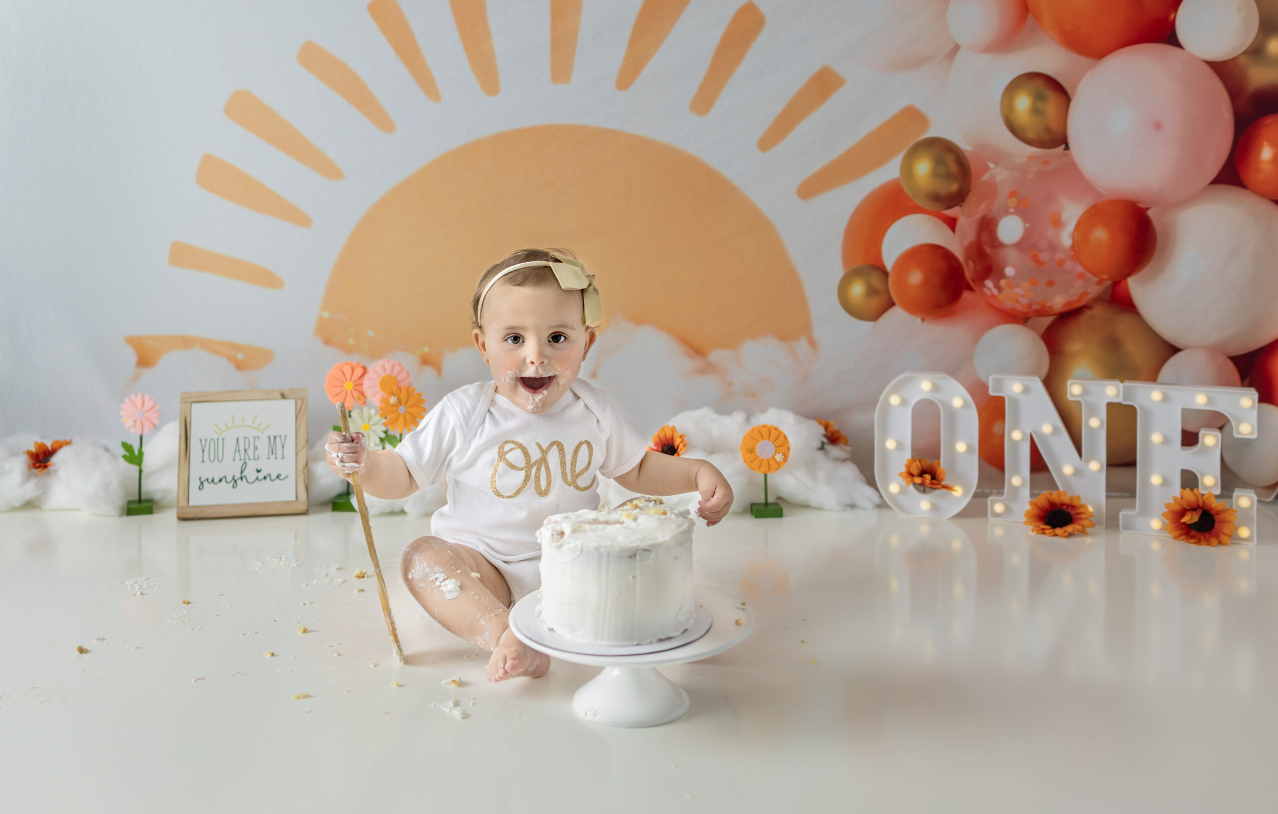 One-year-old girl excitedly digs into birthday cake during sunshine-themed cake smash session.