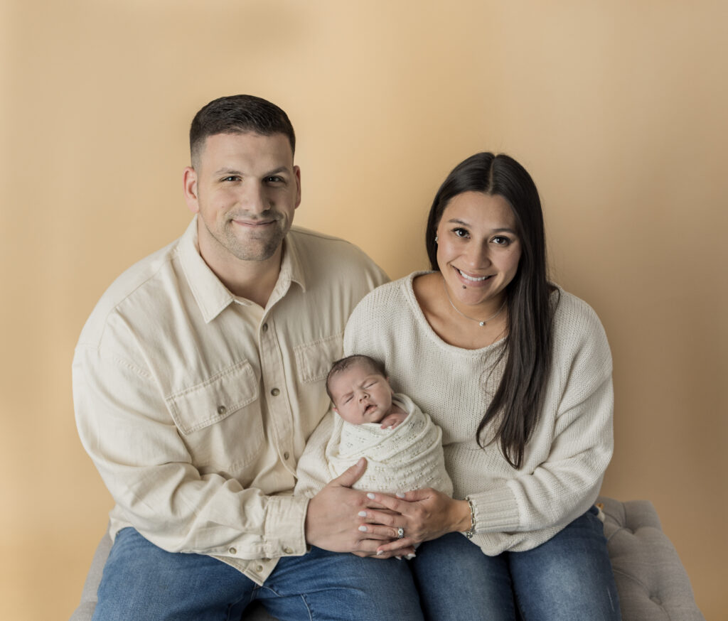 Mom and dad smile while holding their wrapped newborn baby in a warm studio portrait.
