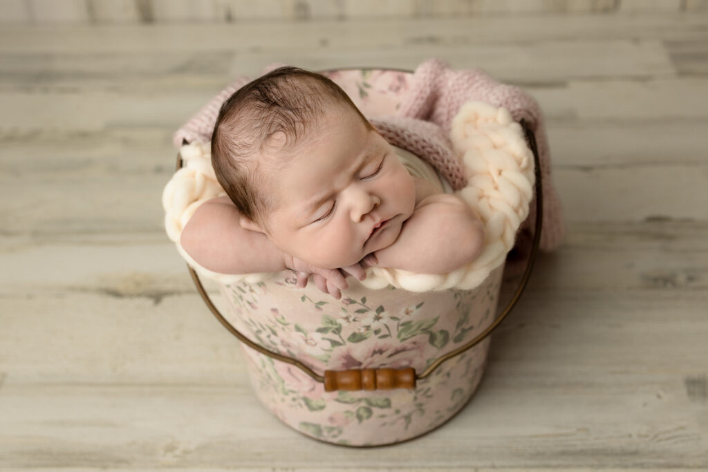 Newborn baby rests with chin on hands inside a pink floral bucket setup during portraits.