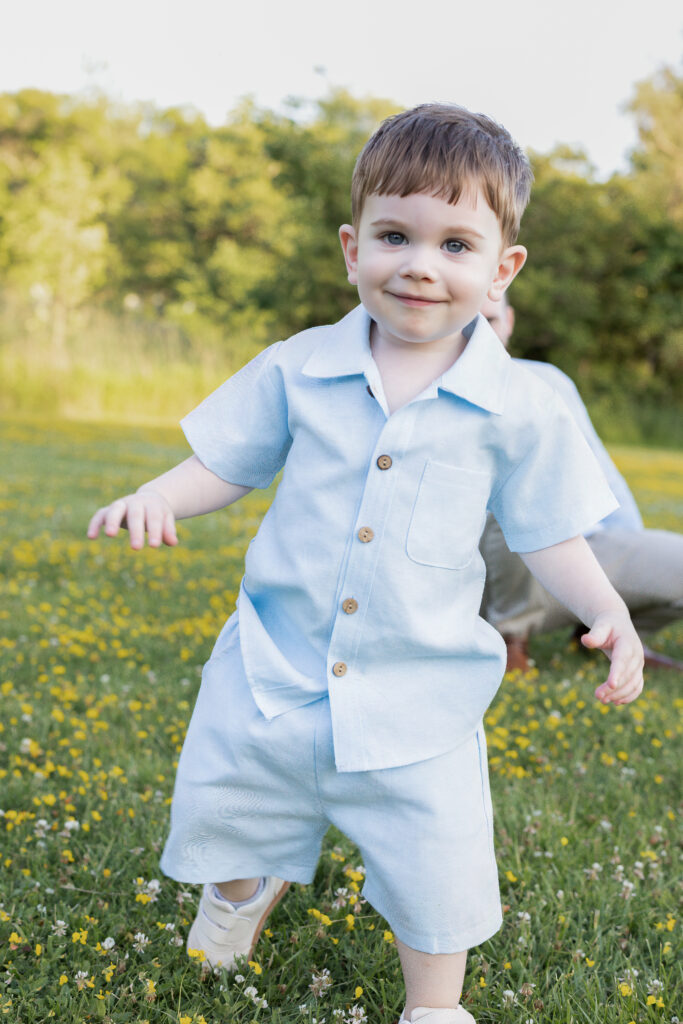 Smiling toddler walks through a grassy summer field filled with tiny yellow and white wildflowers.