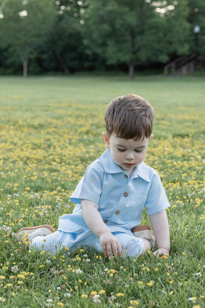 Toddler sits in the grass, looking down at small yellow flowers during an outdoor portrait.