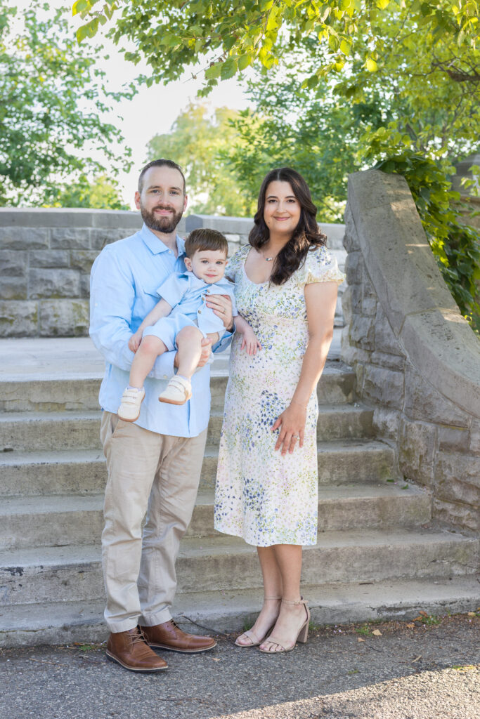 Family of three stands together on stone steps beneath leafy trees in Bergen County, NJ.
