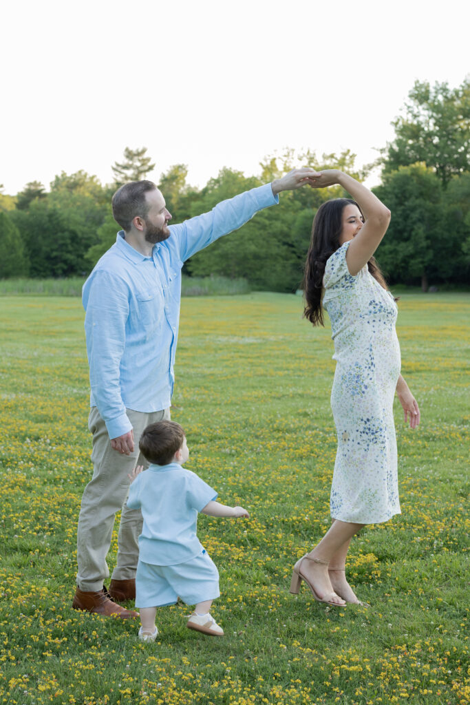 Father twirls mother while their toddler watches during a playful family portrait in the grass.