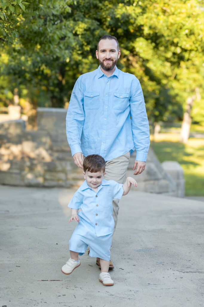 Father walks behind his toddler child on a sunlit stone walkway surrounded by green trees.