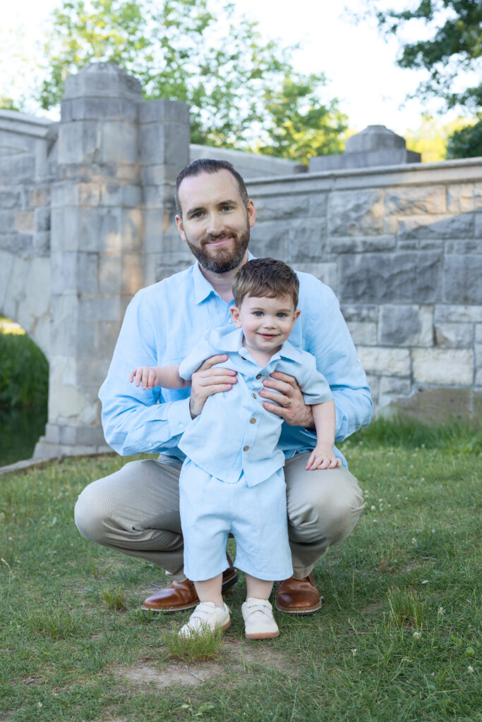 Father kneels behind smiling toddler for a sweet outdoor portrait near a stone bridge wall.