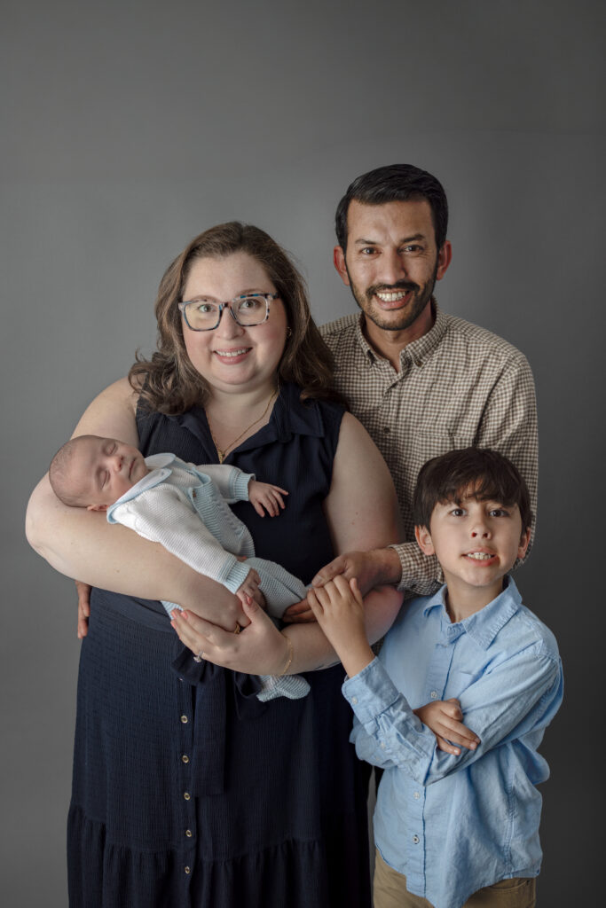 Mother, father, older brother, and newborn baby pose together for a warm family portrait in a studio.