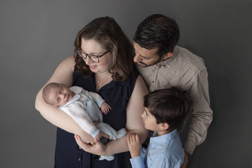 Parents and older brother gently look at their sleeping newborn during a warm and intimate studio family portrait.