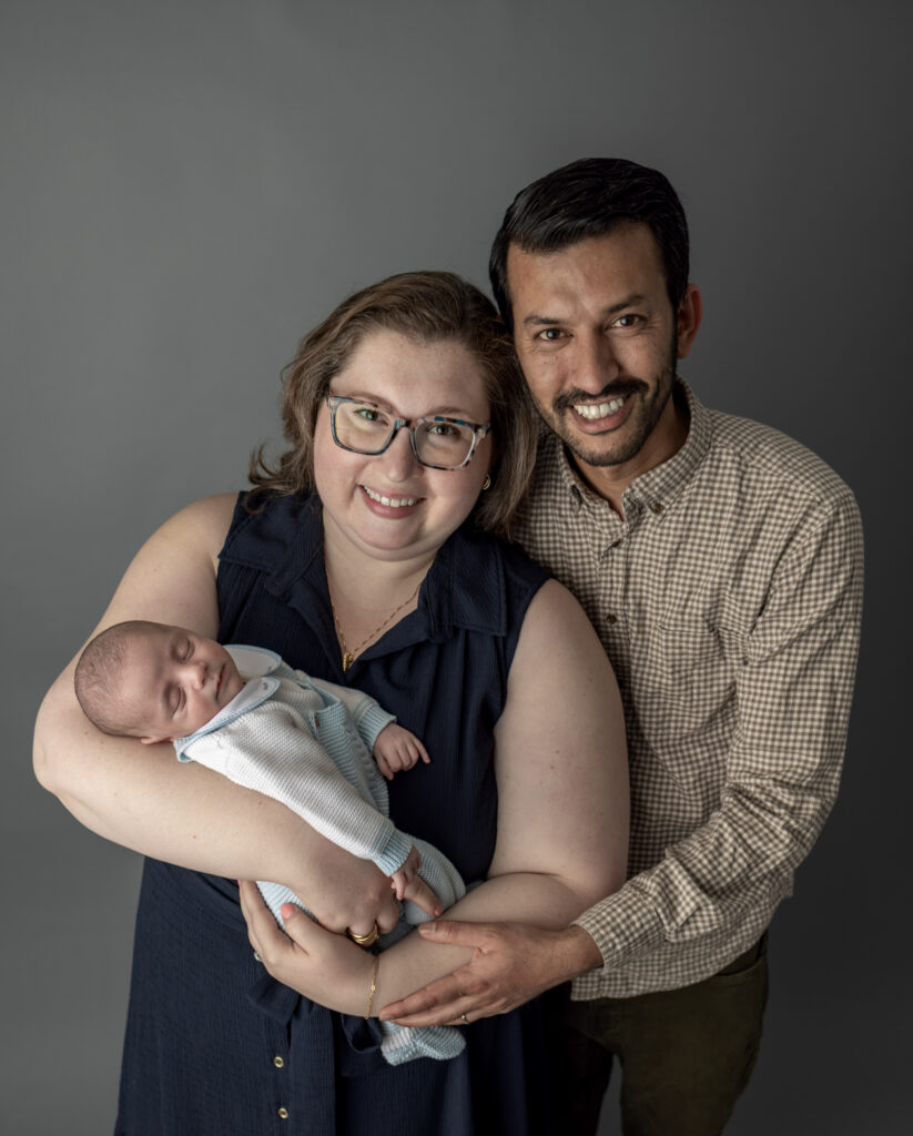 Bergen County, NJ newborn photography session captures parents smiling together while holding their sleeping baby during a portrait.