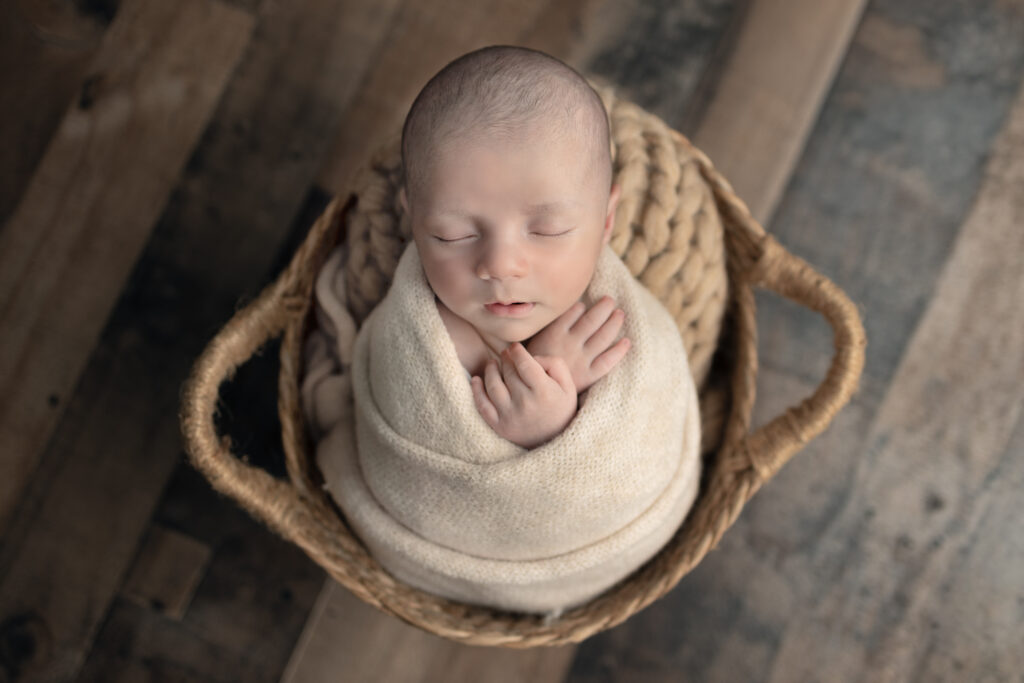 Bergen County, NJ newborn photographer captures sleeping baby wrapped in a soft cream wrap inside a wooden basket.