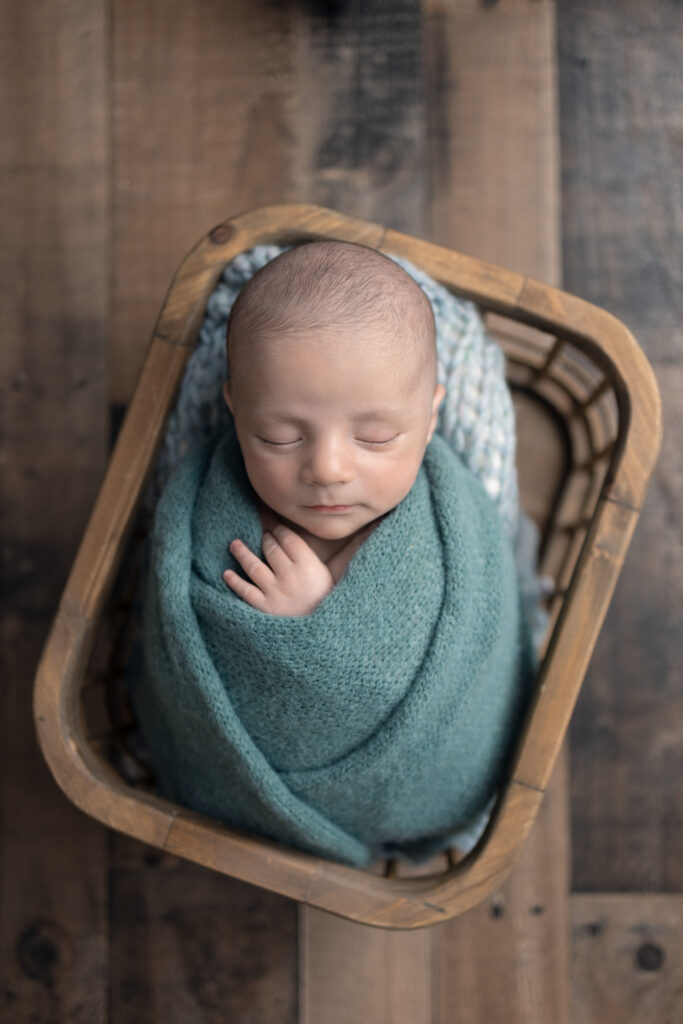 Newborn baby wrapped in a soft green wrap rests peacefully inside a wooden basket during a studio session.
