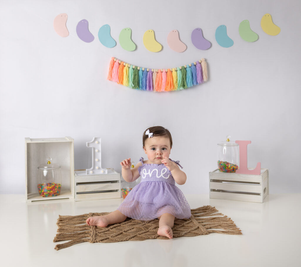 Clifton, NJ cake smash photography featuring a smiling one-year-old girl in a lavender dress seated on a chair.