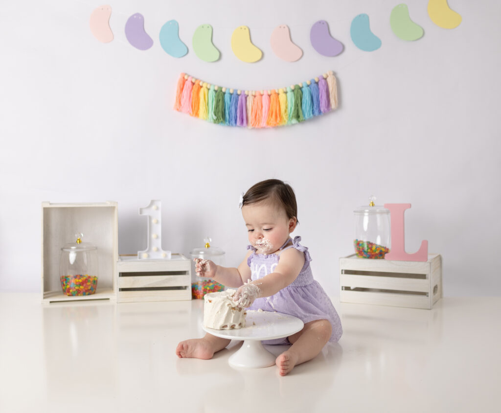 Clifton, NJ first birthday photography features a baby girl enjoying cake with frosting covering her hands and mouth.