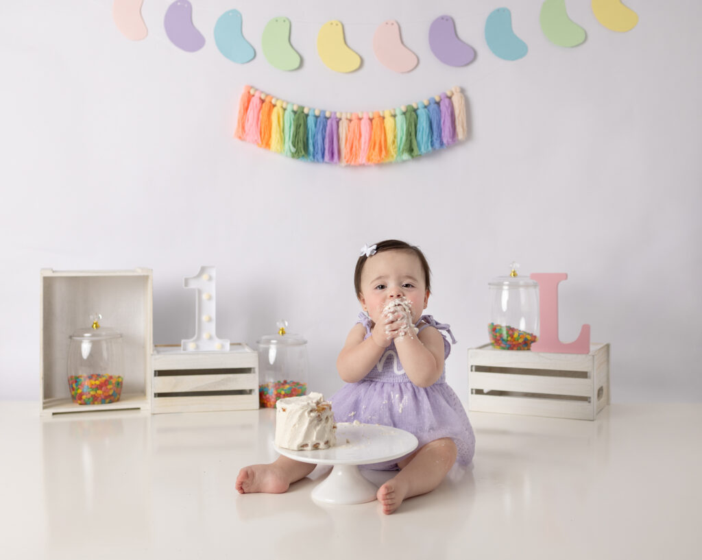 Clifton, NJ cake smash photographer captures a baby girl beside her smashed birthday cake with frosting-covered hands.
