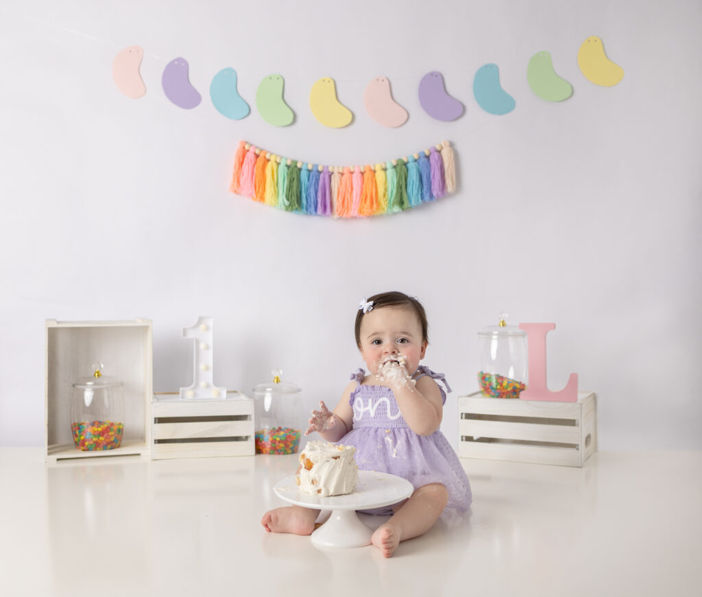 A one-year-old girl happily reaches for her birthday cake during a playful colorful cake smash photography session.