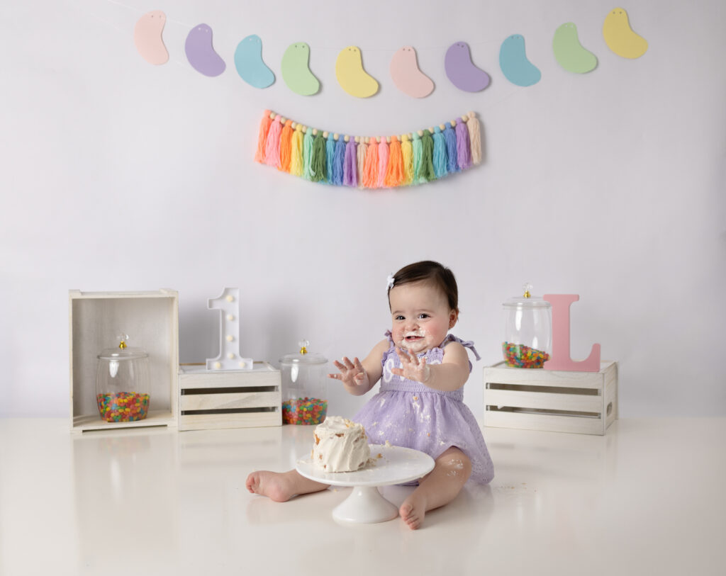 Baby girl sits beside her cake with frosting on her face during a sweet first birthday photography session.