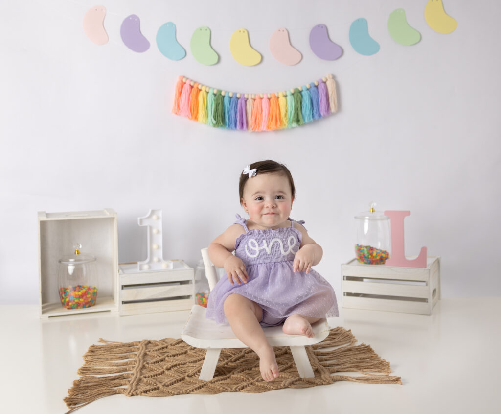 Clifton, NJ cake smash photographer captures a one-year-old girl smiling beneath colorful rainbow decorations in her studio.
