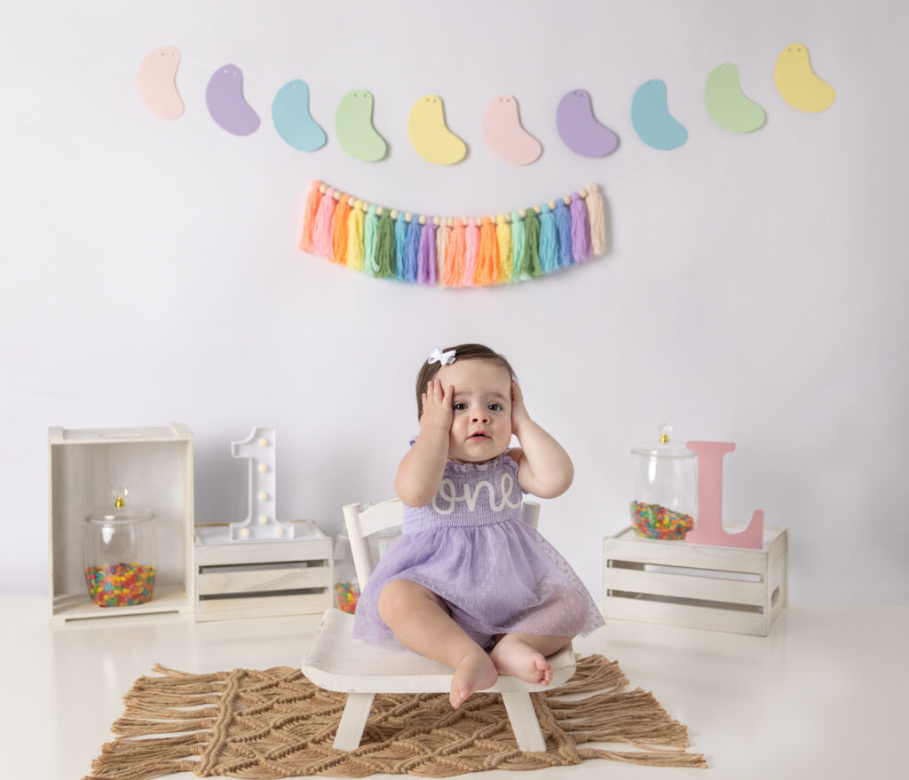 One-year-old girl in a lavender dress sits beneath rainbow decorations, covering her ears during her cake smash session.