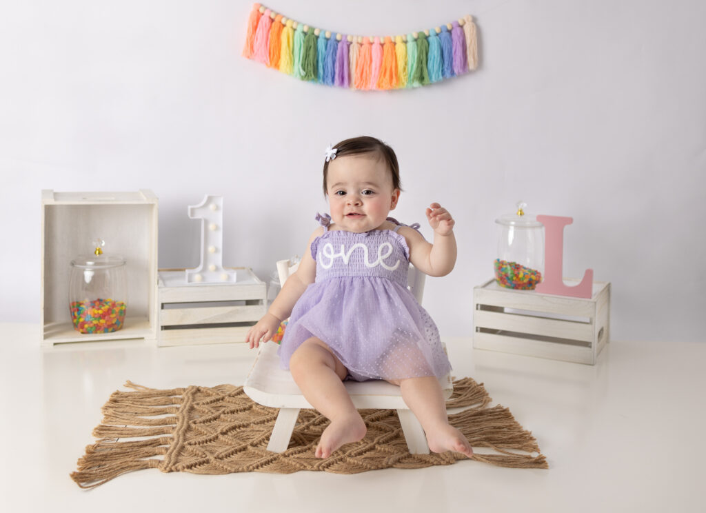 Clifton, NJ first birthday photos feature a baby girl smiling in a lavender dress on a white chair.