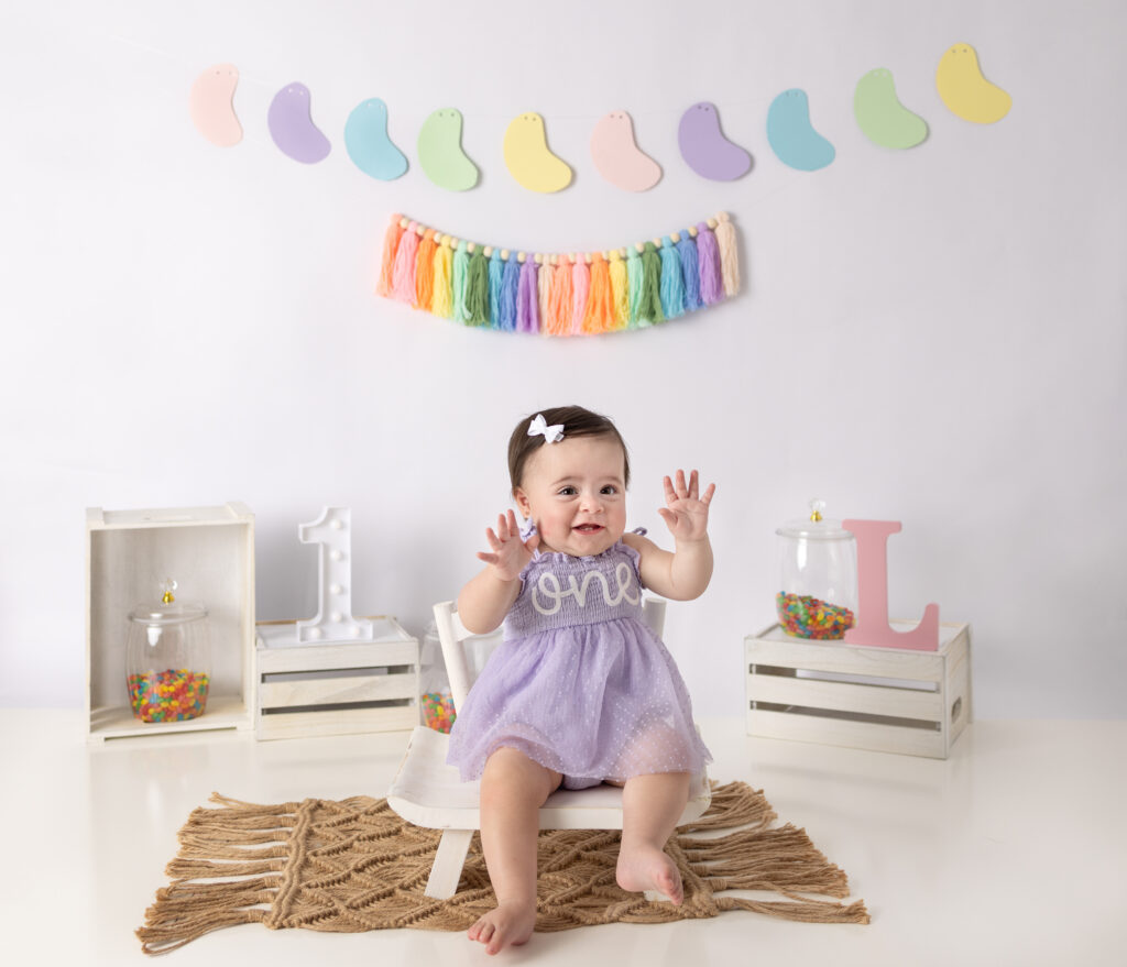 One-year-old girl raises her hands excitedly during a colorful cake smash photography session with rainbow decorations.