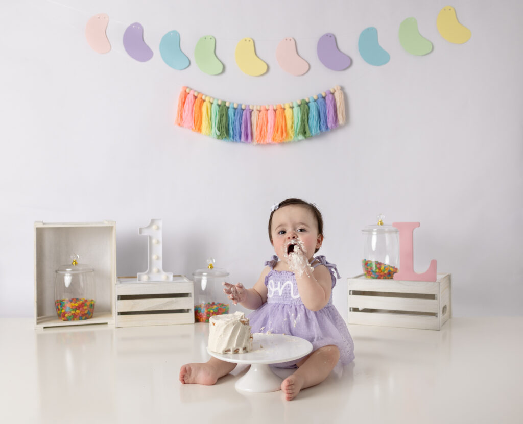 A one-year-old girl explores her birthday cake with frosting-covered hands during a colorful studio cake smash session.