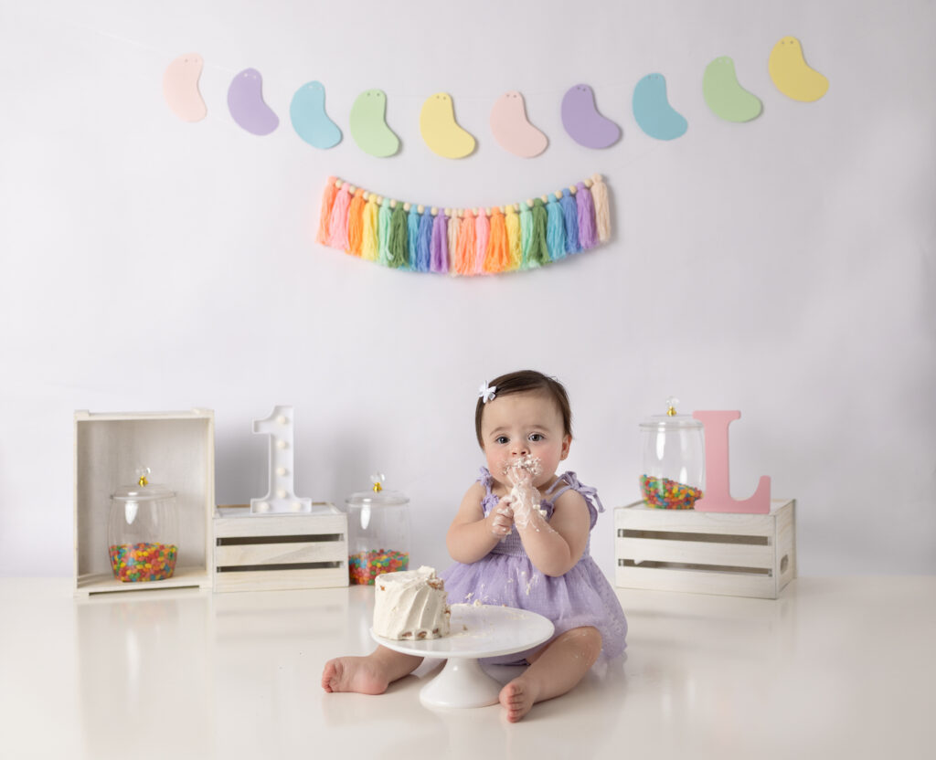 Clifton, NJ cake smash photography captures a baby girl smiling with frosting on her face and hands.