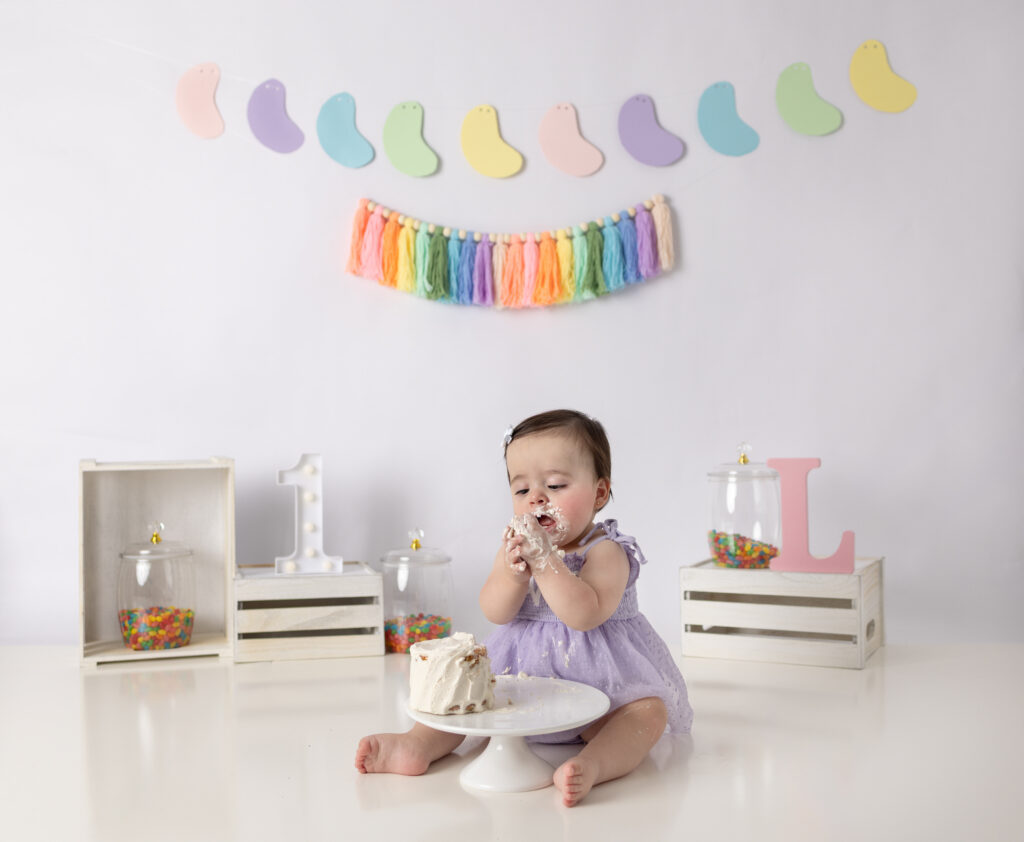 A happy one-year-old girl reaches toward her cake during a playful lavender-themed first birthday photography session.